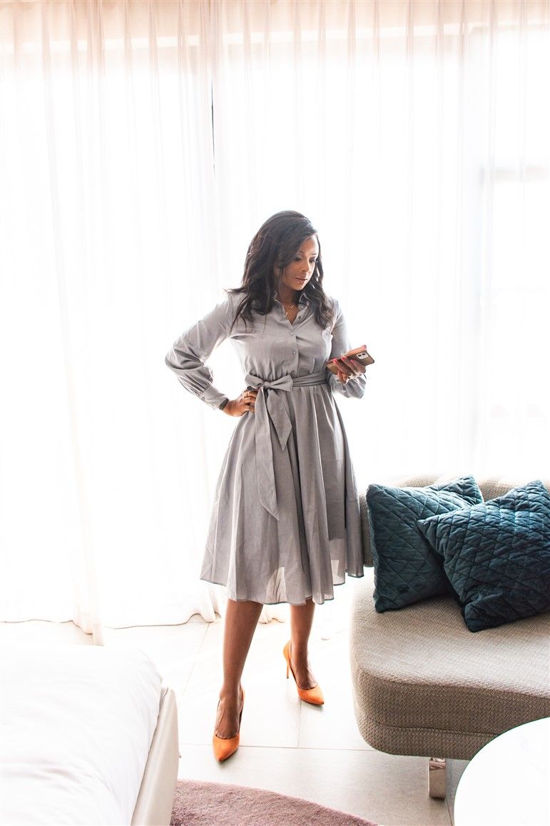 Woman wearing gray belted dress and heels posing in bright interior setting.