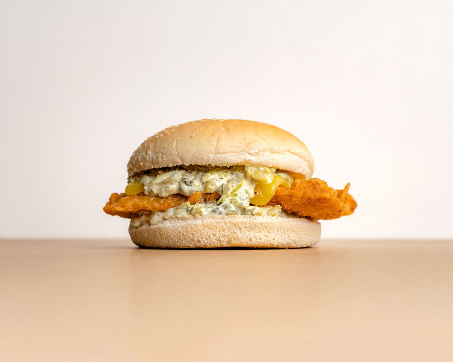 Crispy fish sandwich with tartar sauce on plain bun against white background.