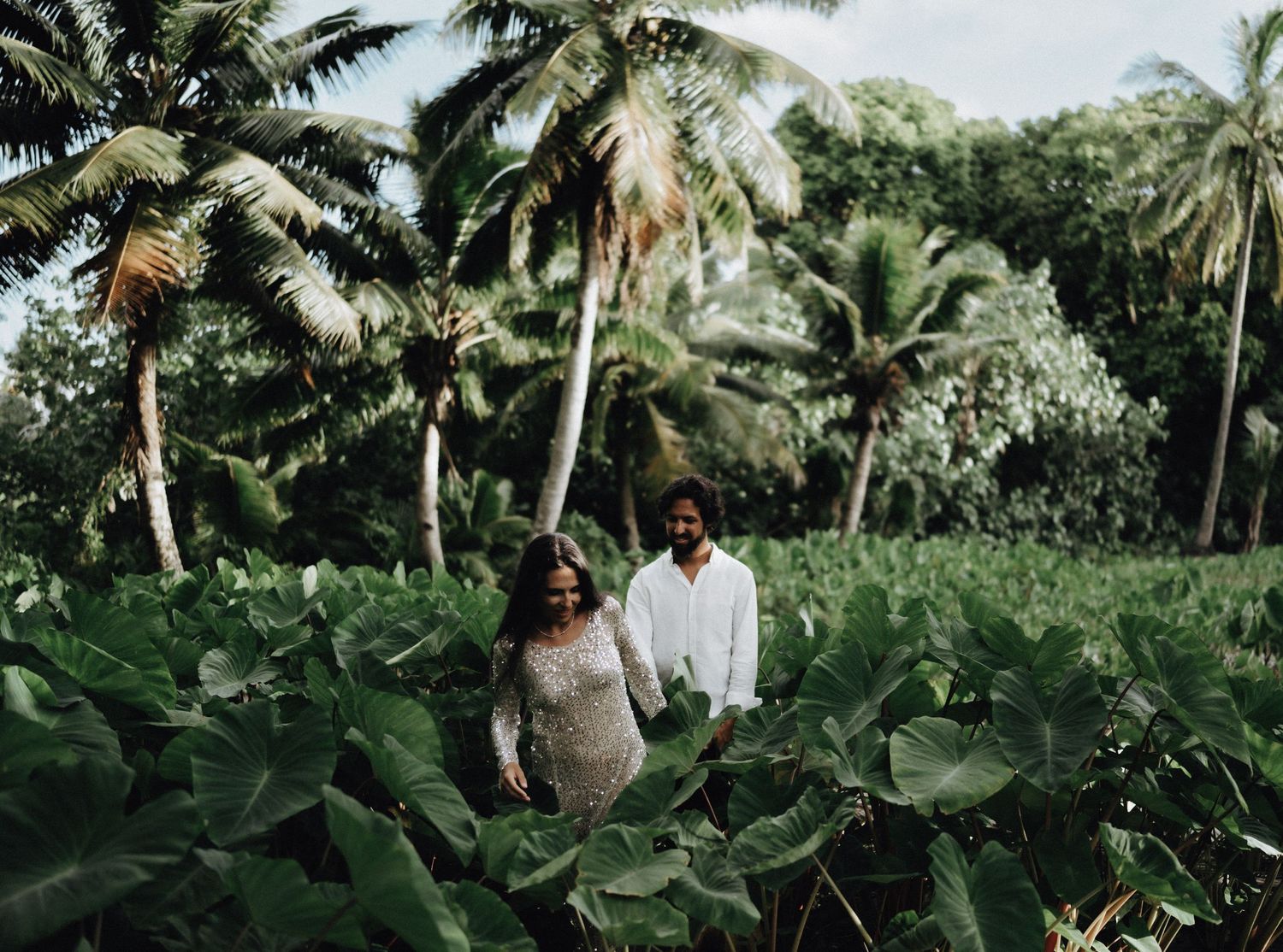 A couple stands among lush tropical foliage and palm trees in a garden setting.