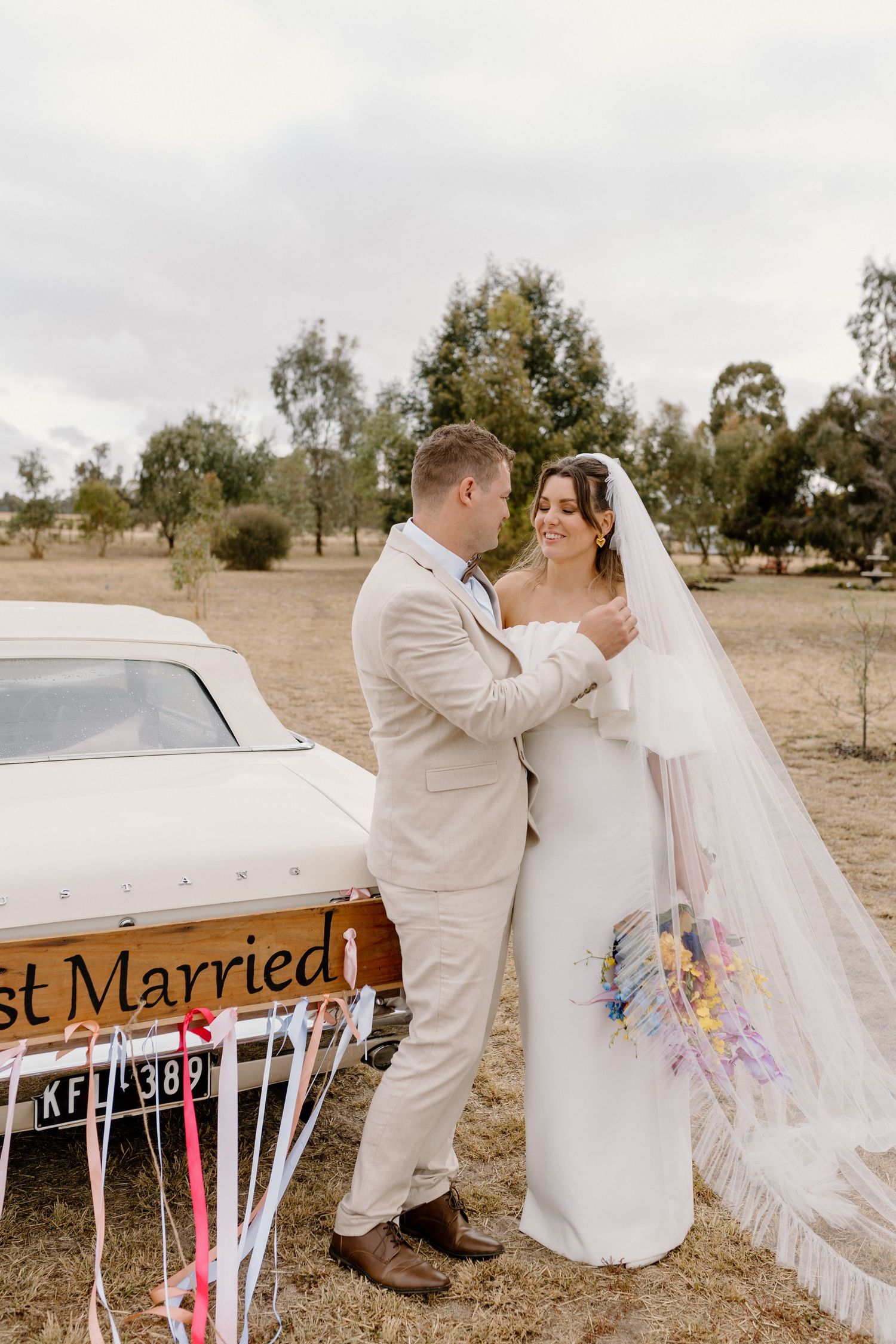 Newlyweds embrace by vintage car with 'Just Married' sign in rustic countryside setting.