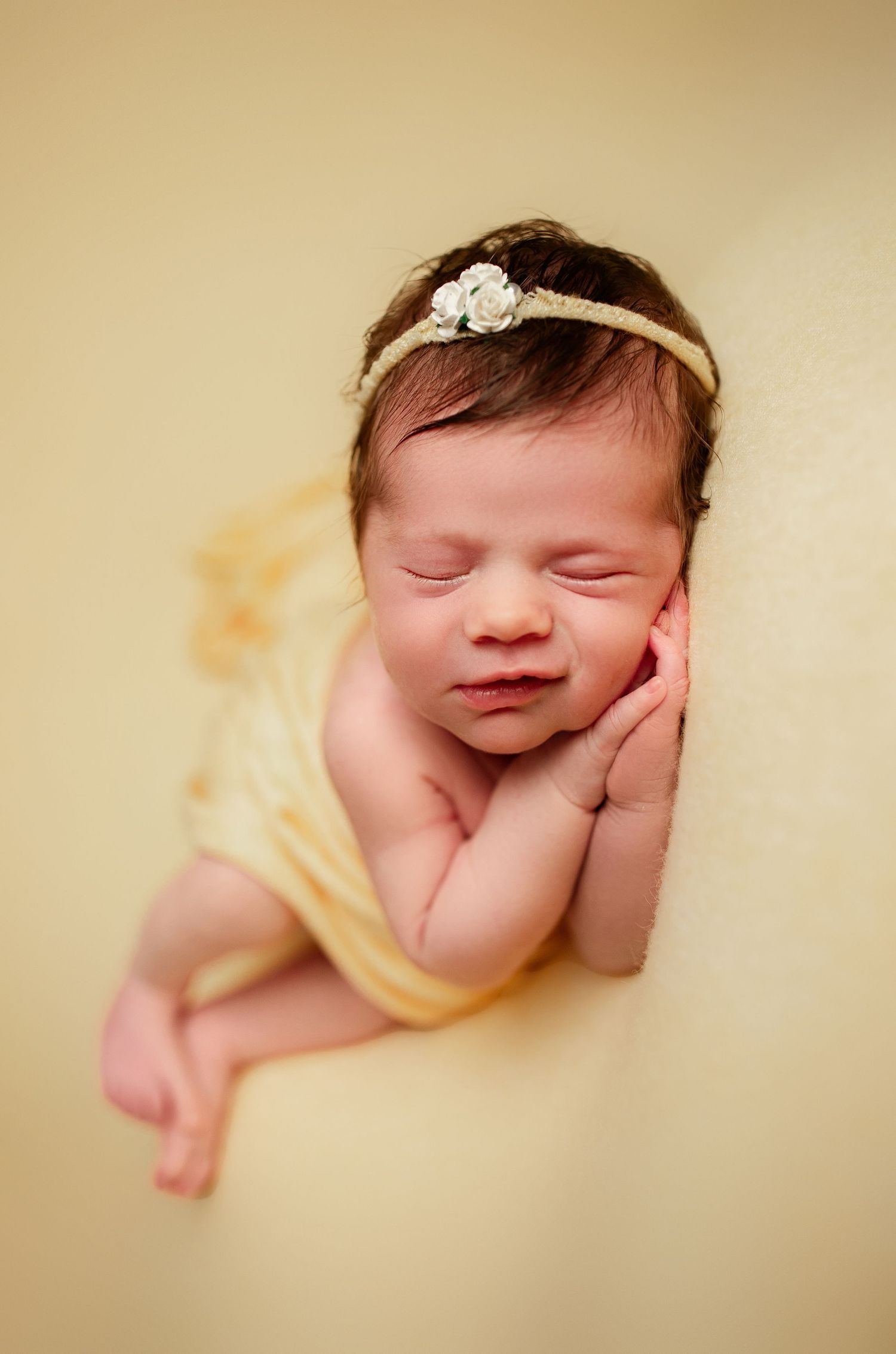 Sleeping newborn wearing delicate floral headband wrapped in yellow fabric against cream backdrop.