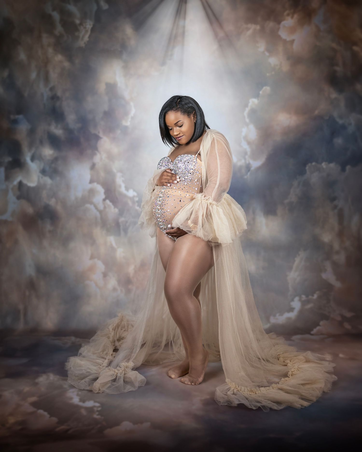 Ethereal maternity photo with flowing cream dress and dramatic cloudy background with glowing light.