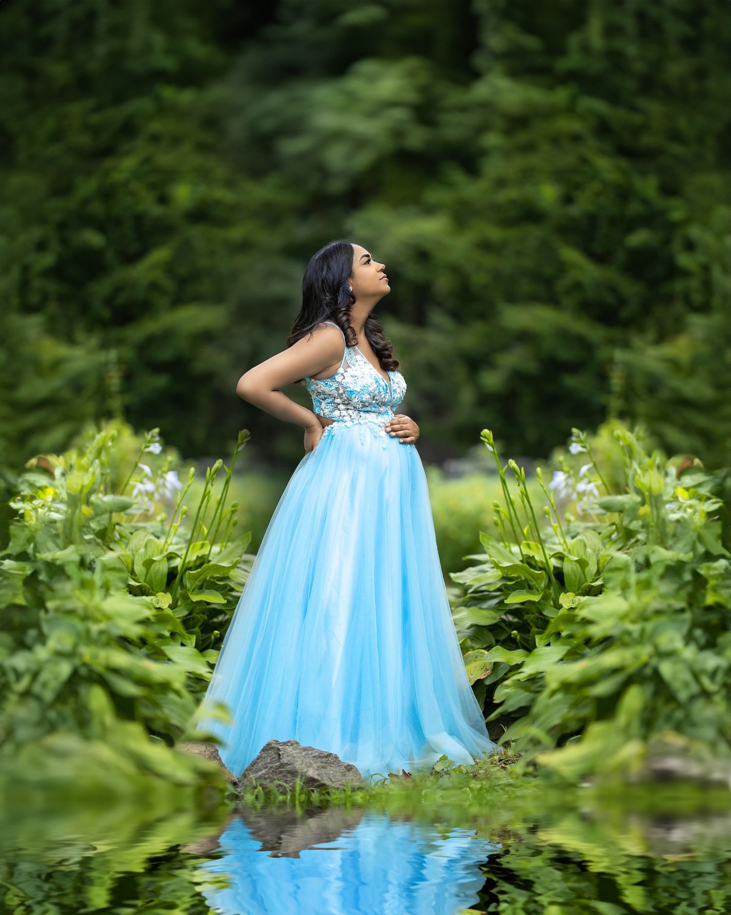 Ethereal maternity portrait in blue gown reflecting in garden pond surrounded by greenery.