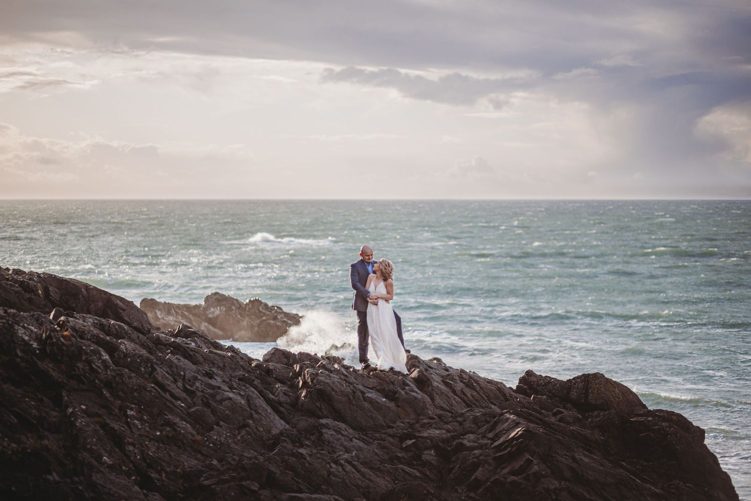 A bride and groom stand on a rocky cliff at their ocean side elopement
