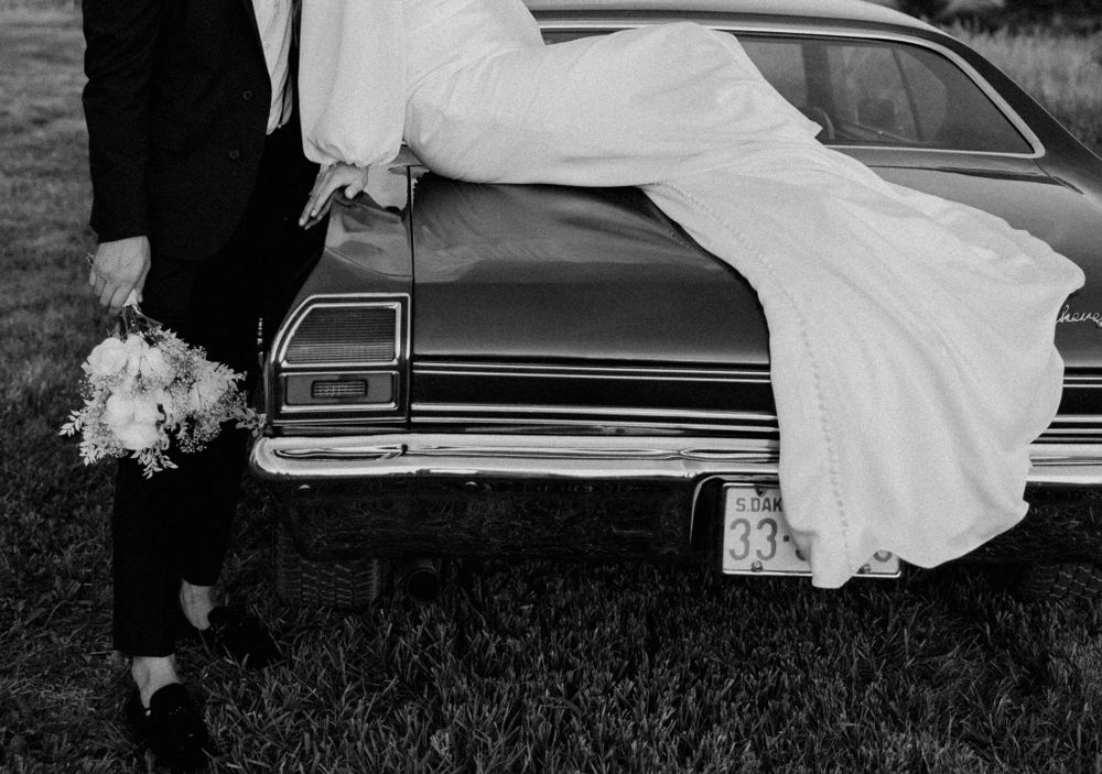 Black and white photo of newlyweds posing with a vintage car, showing dress and bouquet details.