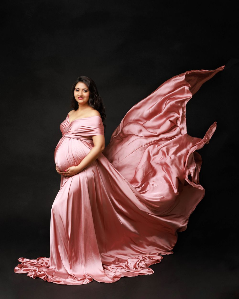 Flowing pink satin maternity gown with dramatic fabric movement against dark background.