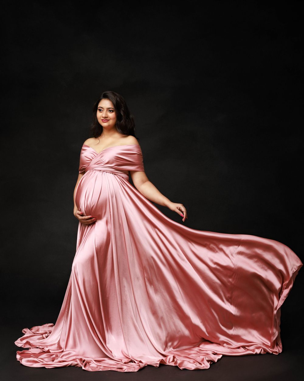 Dramatic maternity portrait in flowing pink satin gown against dark backdrop.