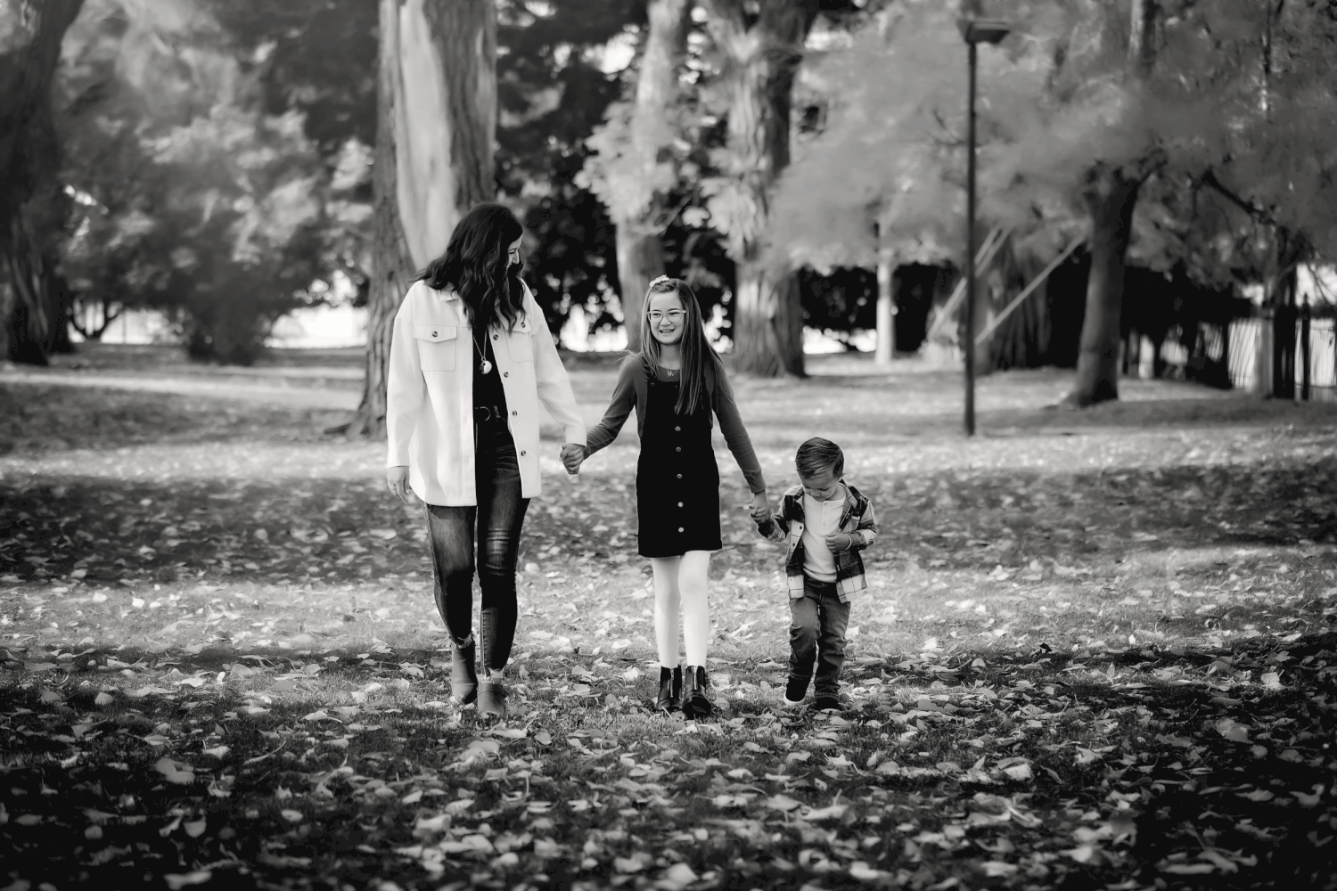 A black and white photograph shows three people walking together through a park scattered with fallen leaves.