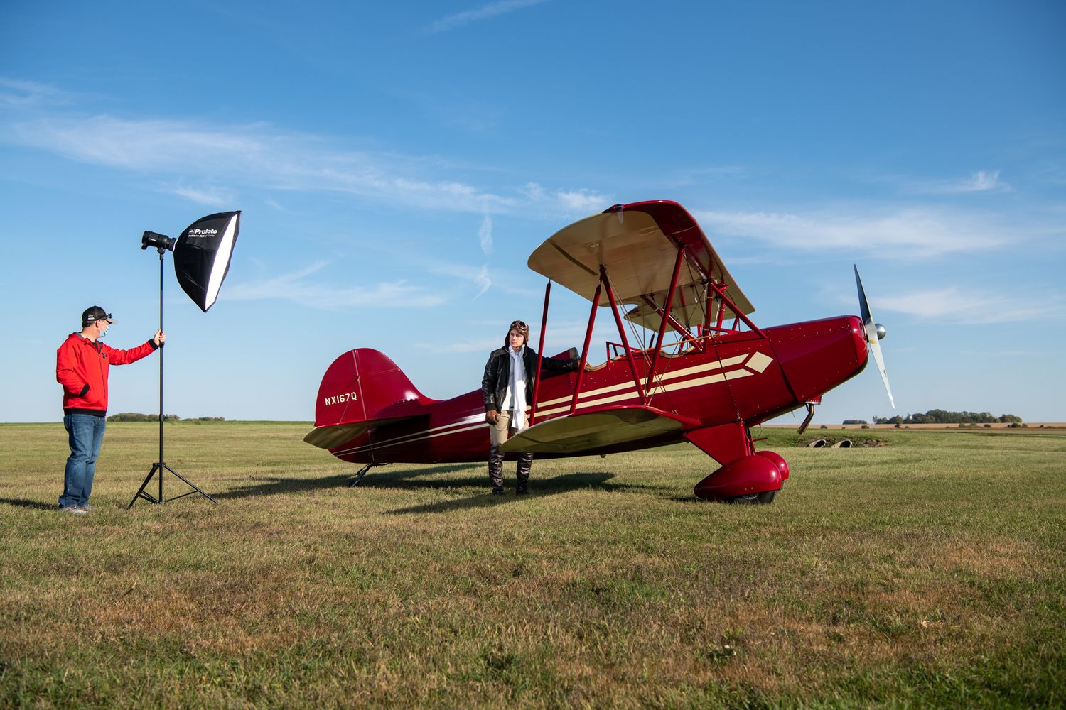 A vintage red biplane parked on grass field while photographer sets up lighting equipment against blue sky.