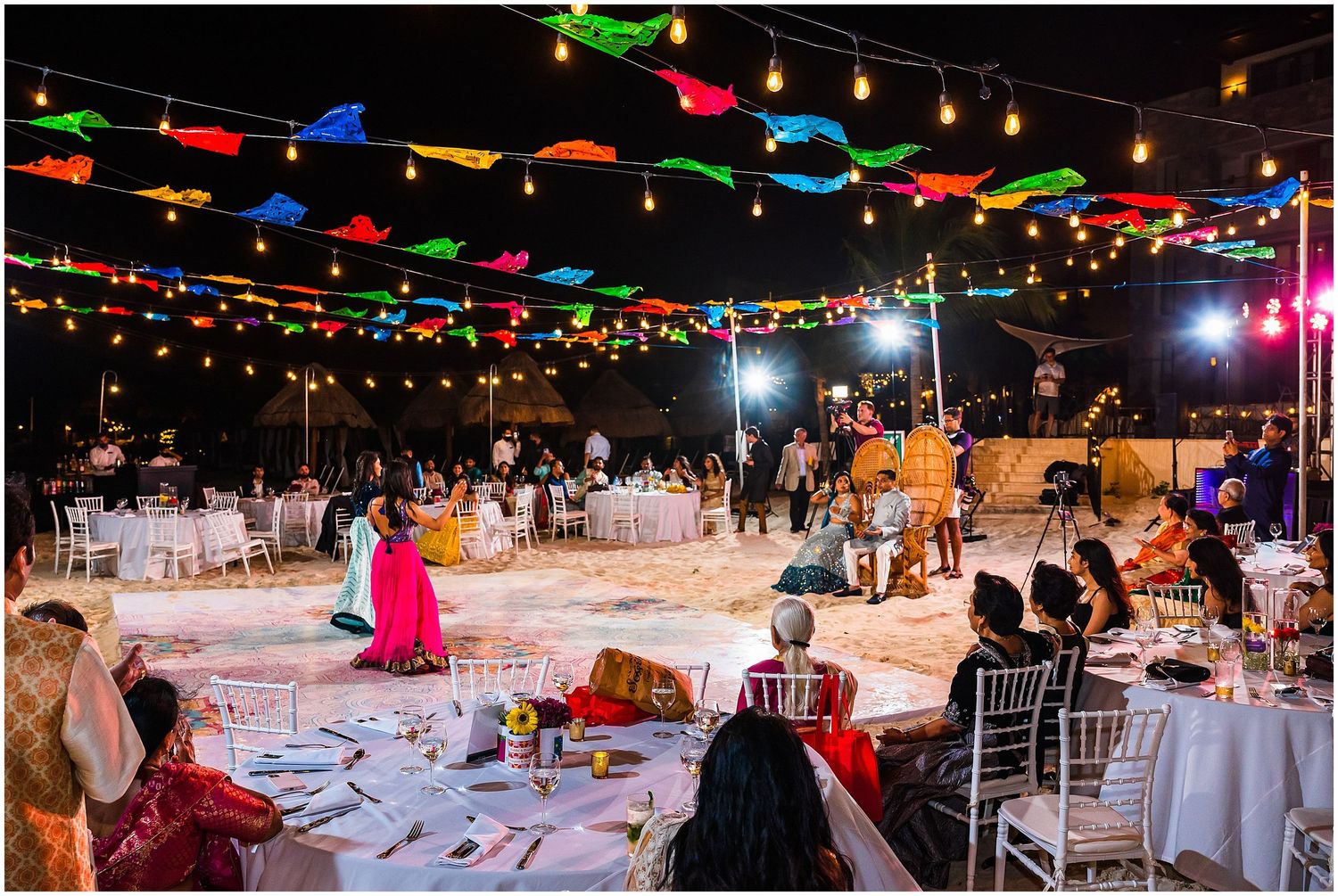 Beach wedding reception under colorful string lights with round tables and chairs.