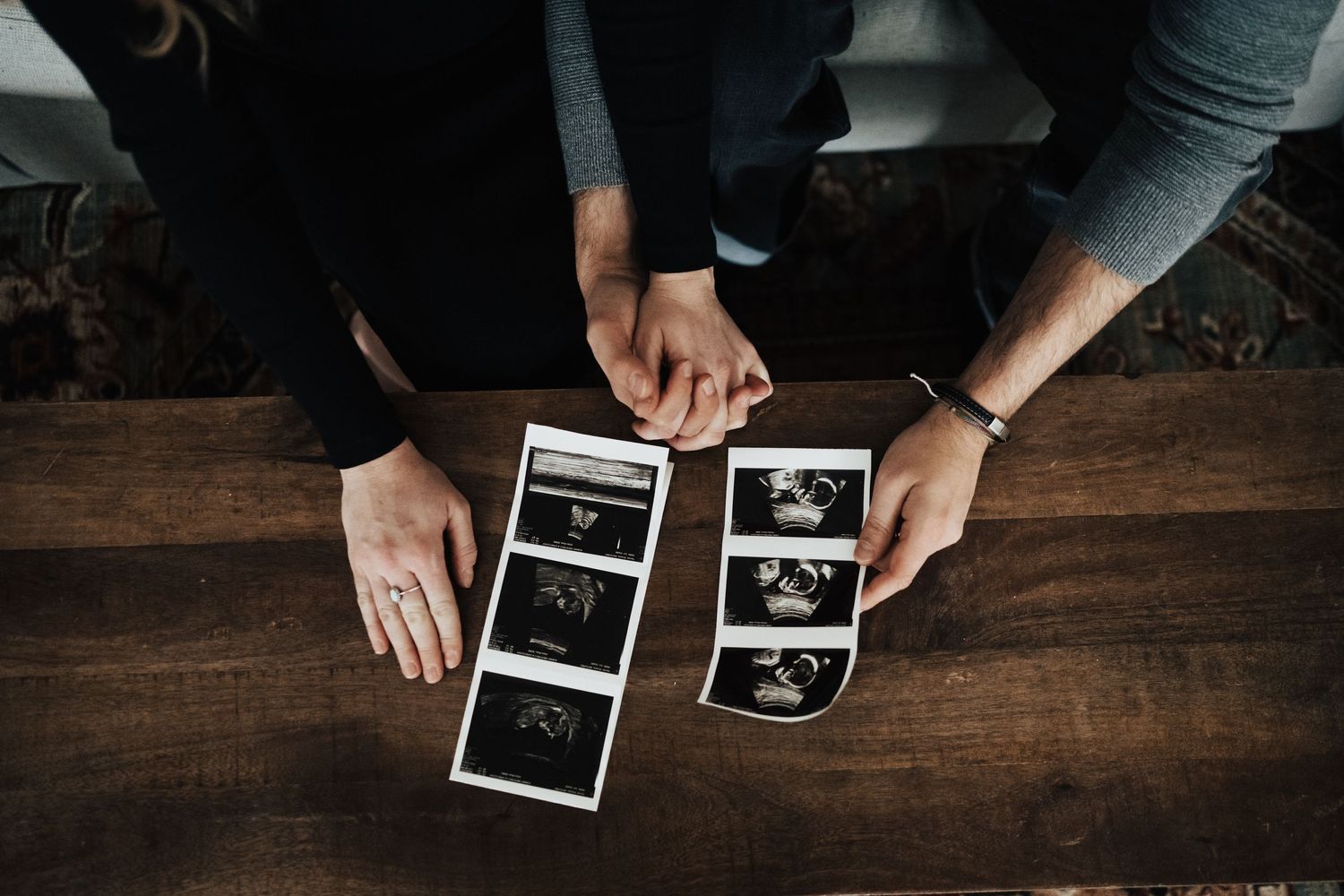 Couple holds hands while viewing ultrasound photos on a wooden table during pregnancy.