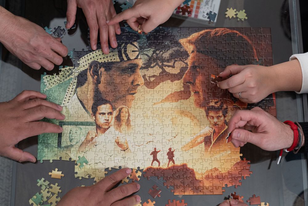 Multiple hands working together to complete a movie-themed jigsaw puzzle on a dark surface.