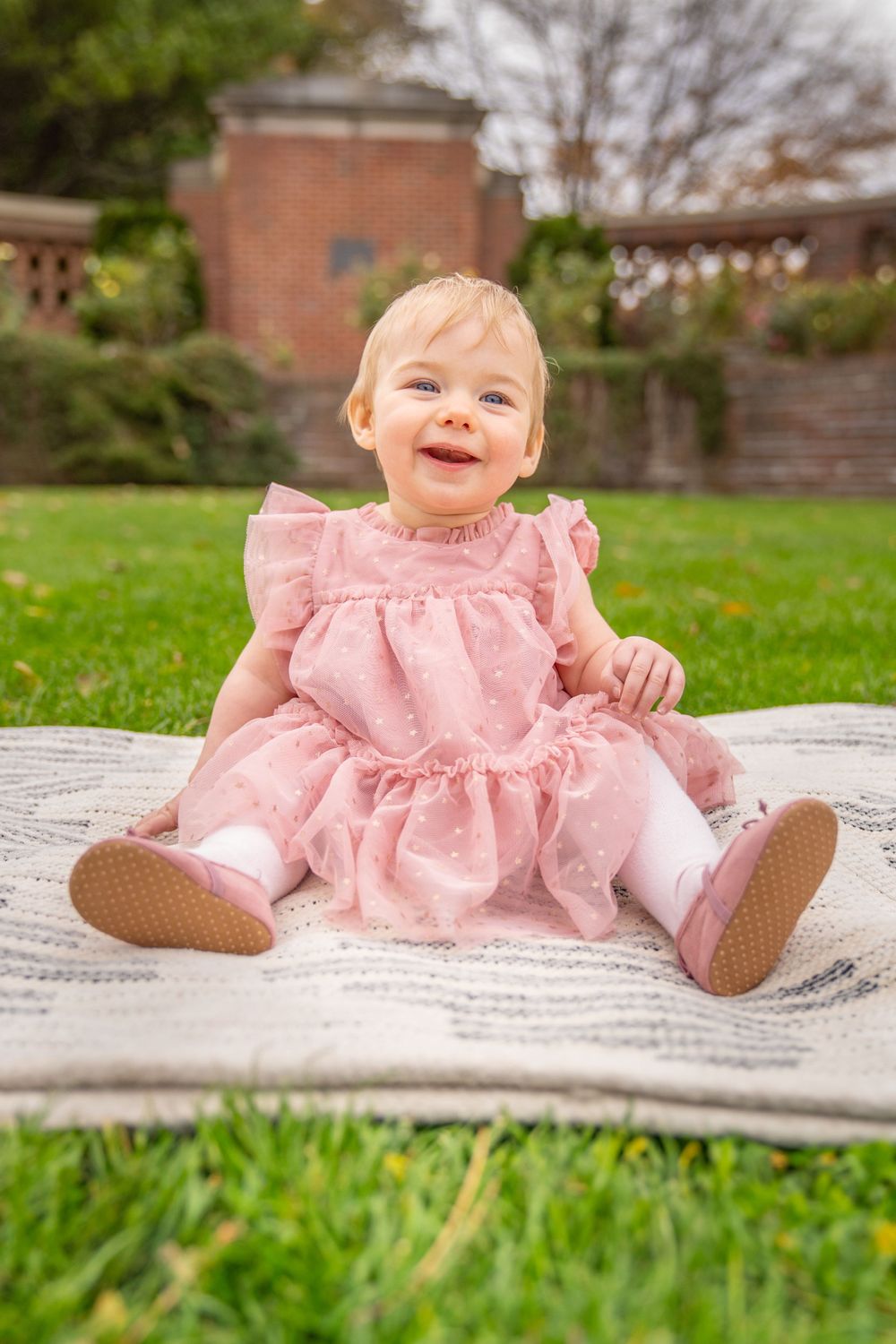Baby wearing pink ruffled dress sitting on blanket in garden.