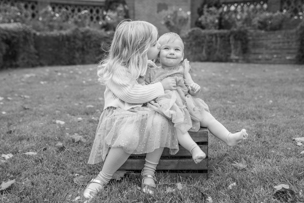 Two children in light dresses sitting together on wooden swing in garden setting.
