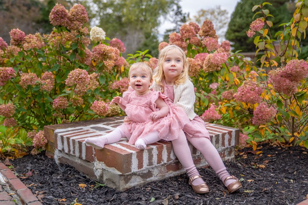 Two siblings sitting together on brick steps surrounded by pink hydrangea bushes.
