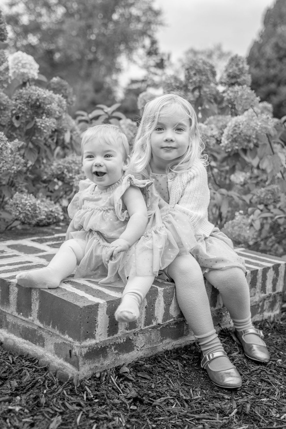 Black and white portrait of two young children sitting on garden steps among flowering bushes.