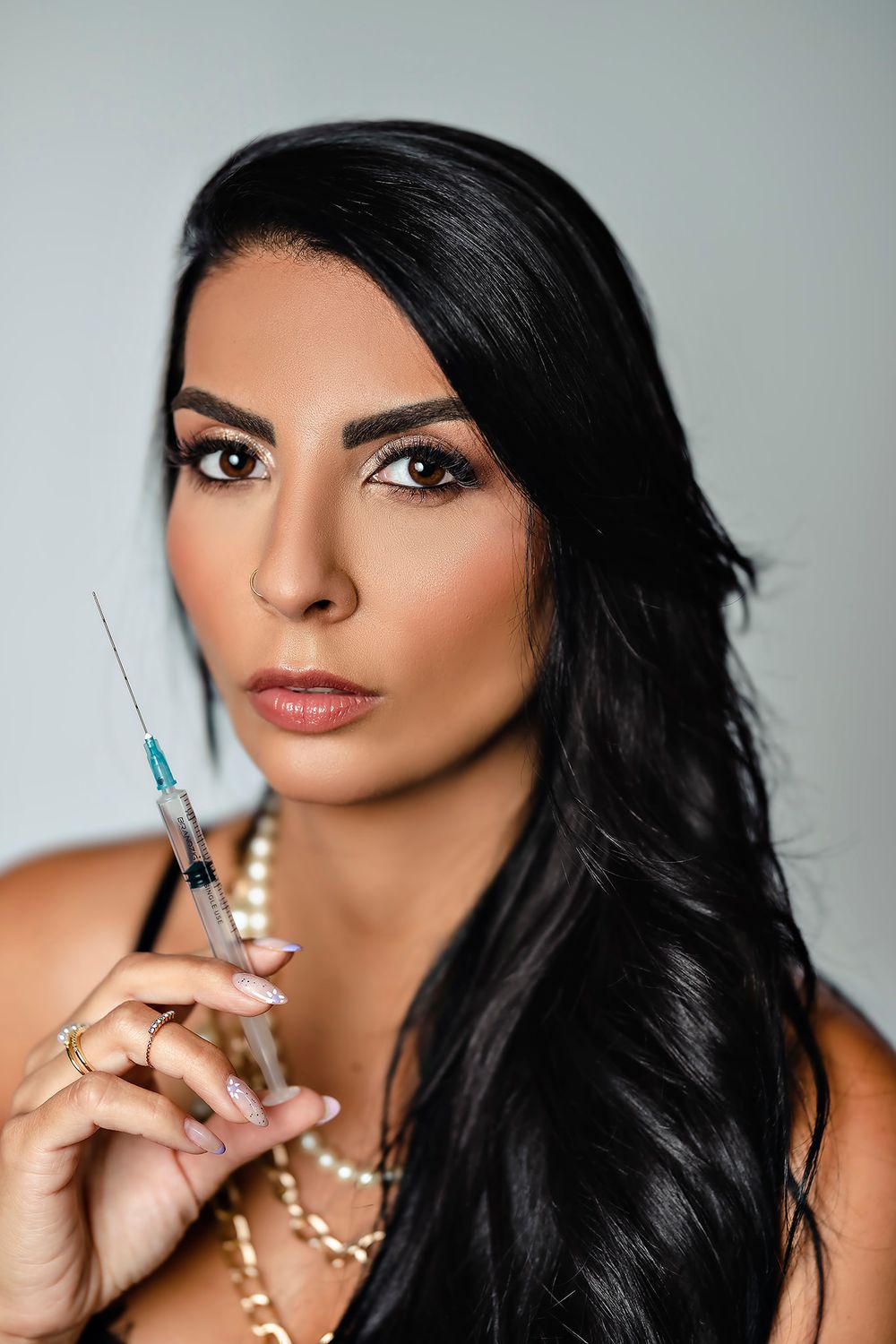 Professional beauty photoshoot featuring syringes and pearl necklace accessories against gray background.