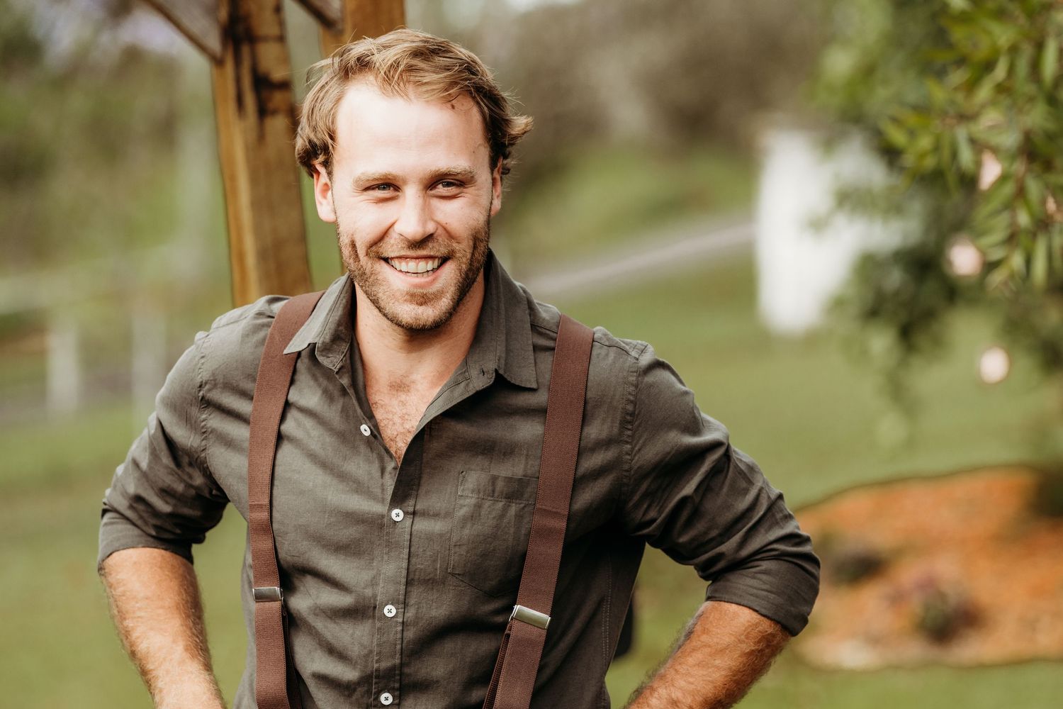 A person wearing suspenders and a button-down shirt smiles outdoors.