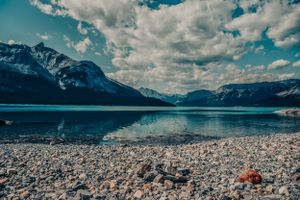 Rocky mountain lake shore with snow-capped peaks and dramatic clouds reflected in clear water.