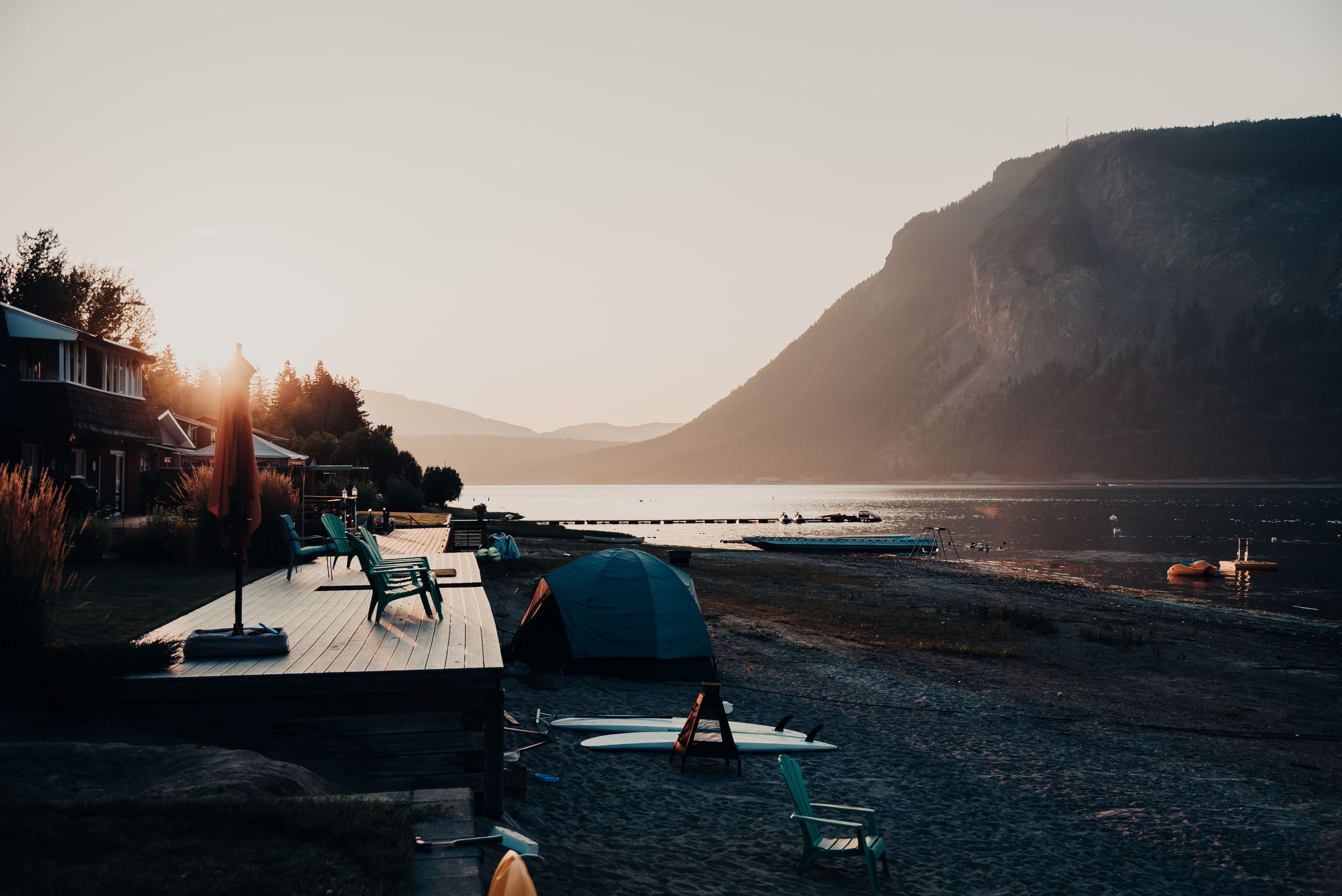 Sunset silhouettes boats and beach furniture along mountain lake shore.