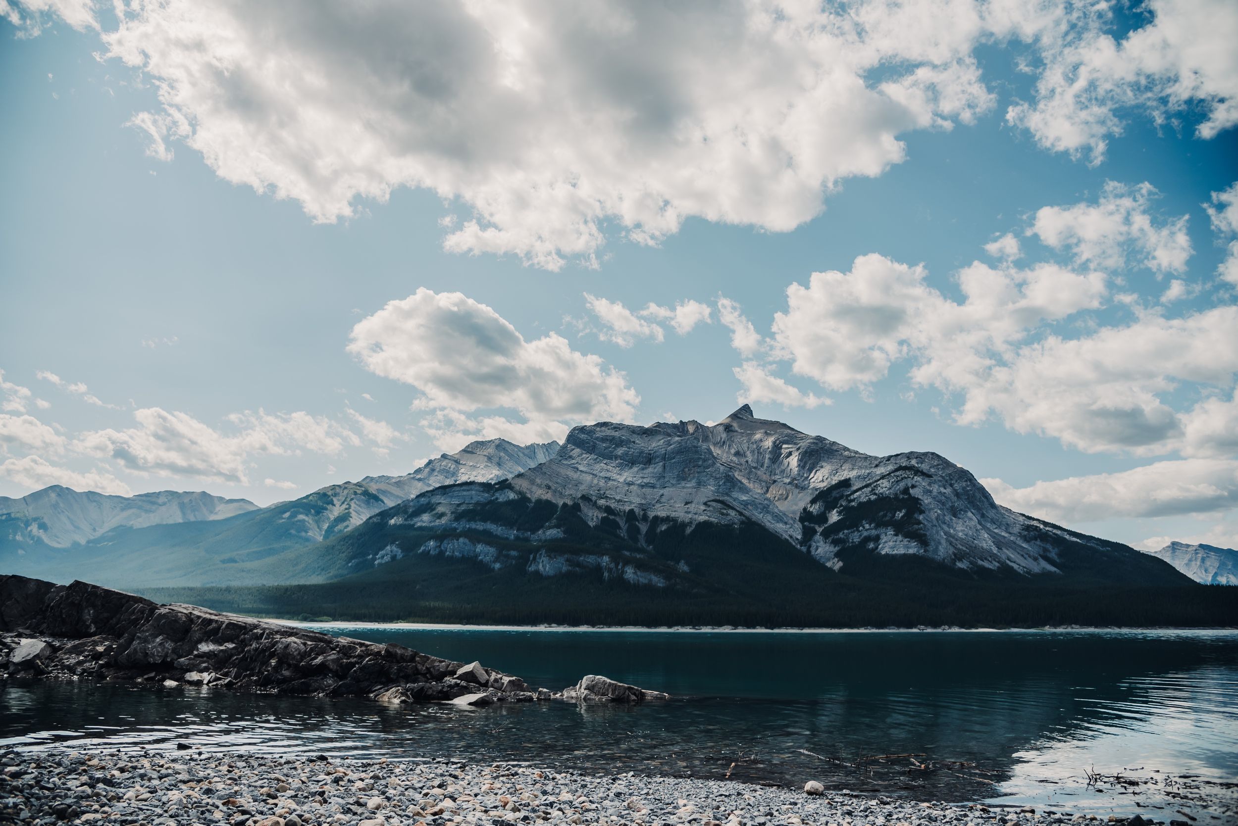 Snow-capped mountain peak reflects in clear lake waters under fluffy white clouds.
