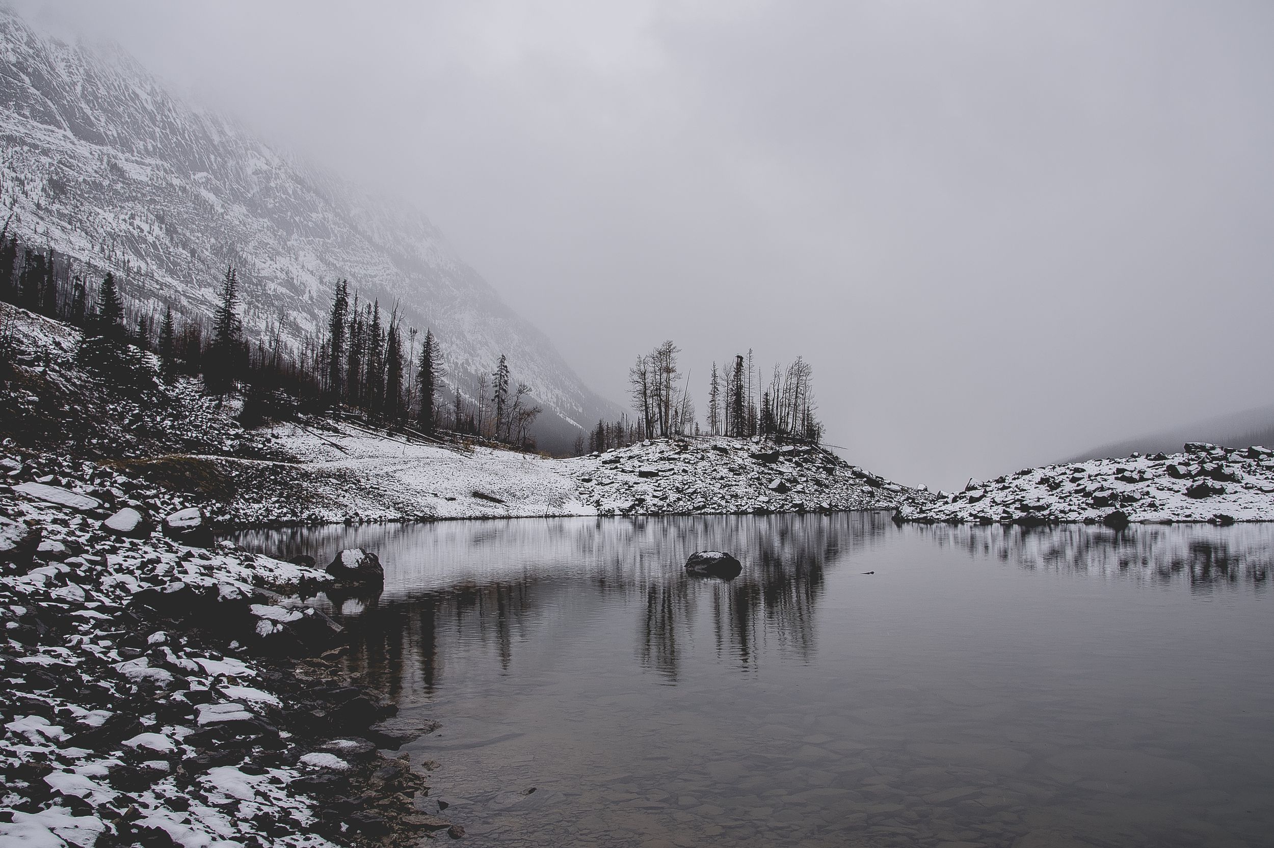 Snow-covered mountain landscape reflects in a calm alpine lake under overcast winter skies.