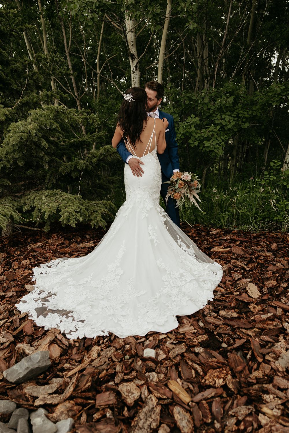 Couple embraces on rocky forest path showing off bride's elegant lace train.