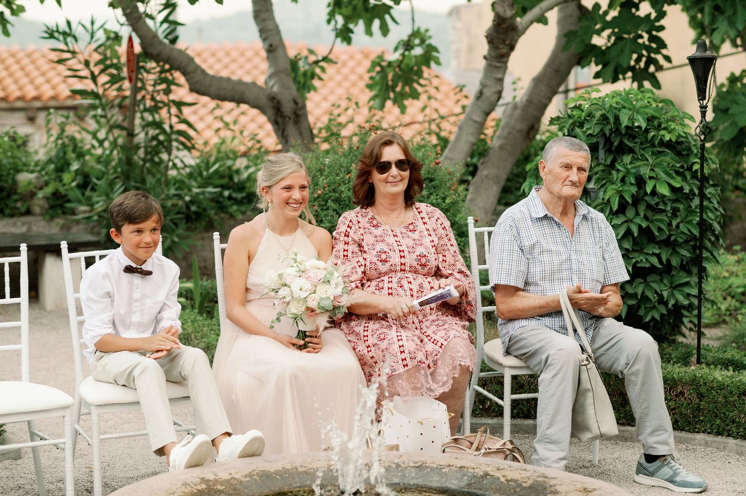 Wedding guests seated in white chairs surrounded by lush greenery in an outdoor garden setting.
