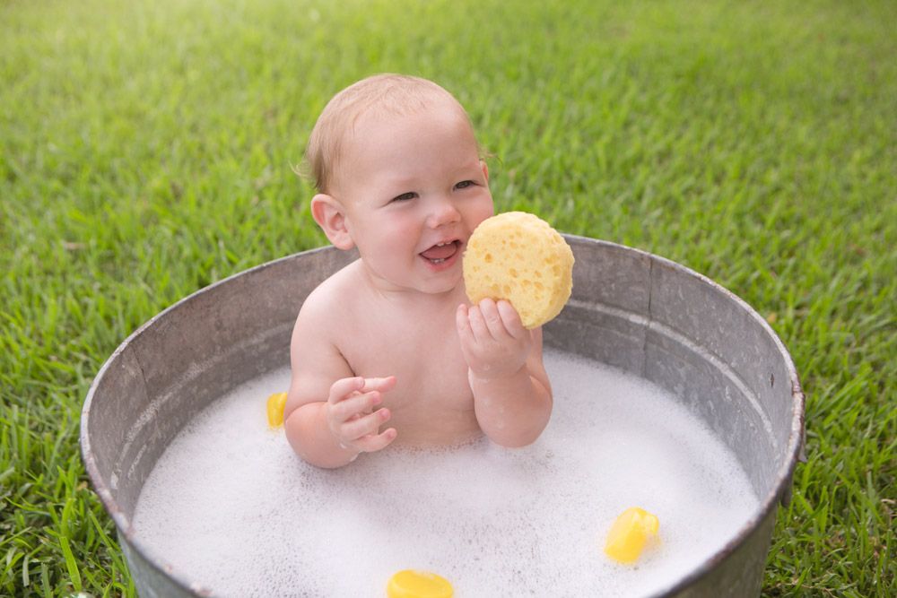 Baby sitting in metal tub with bubbles and yellow sponge on grass.