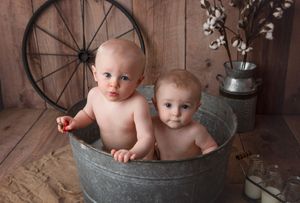 Two babies sitting in rustic metal wash tub with vintage wheel and floral decor in background.