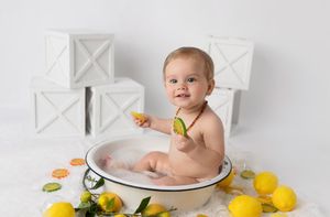 Baby in white tub surrounded by citrus fruits and white decorative boxes.