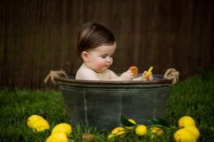 Baby sits in rustic metal tub with rubber ducks on green grass with scattered lemons around.