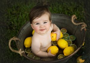 Smiling baby in rustic metal tub surrounded by bright yellow lemons on grass.