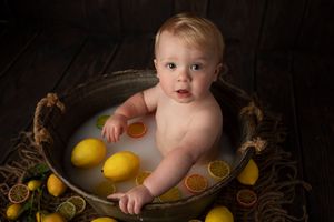 Baby in vintage metal tub with scattered lemons and citrus slices on dark wood background.