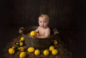 Baby sitting in rustic metal tub surrounded by fresh yellow lemons on wooden floor.
