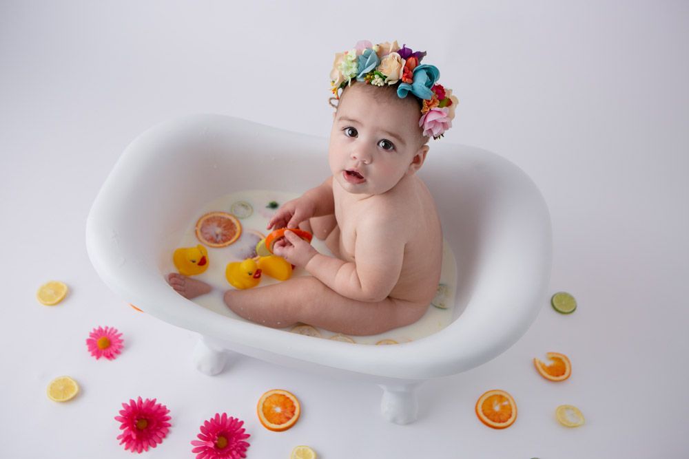 Baby in white bathtub with floral crown and colorful flower decorations.