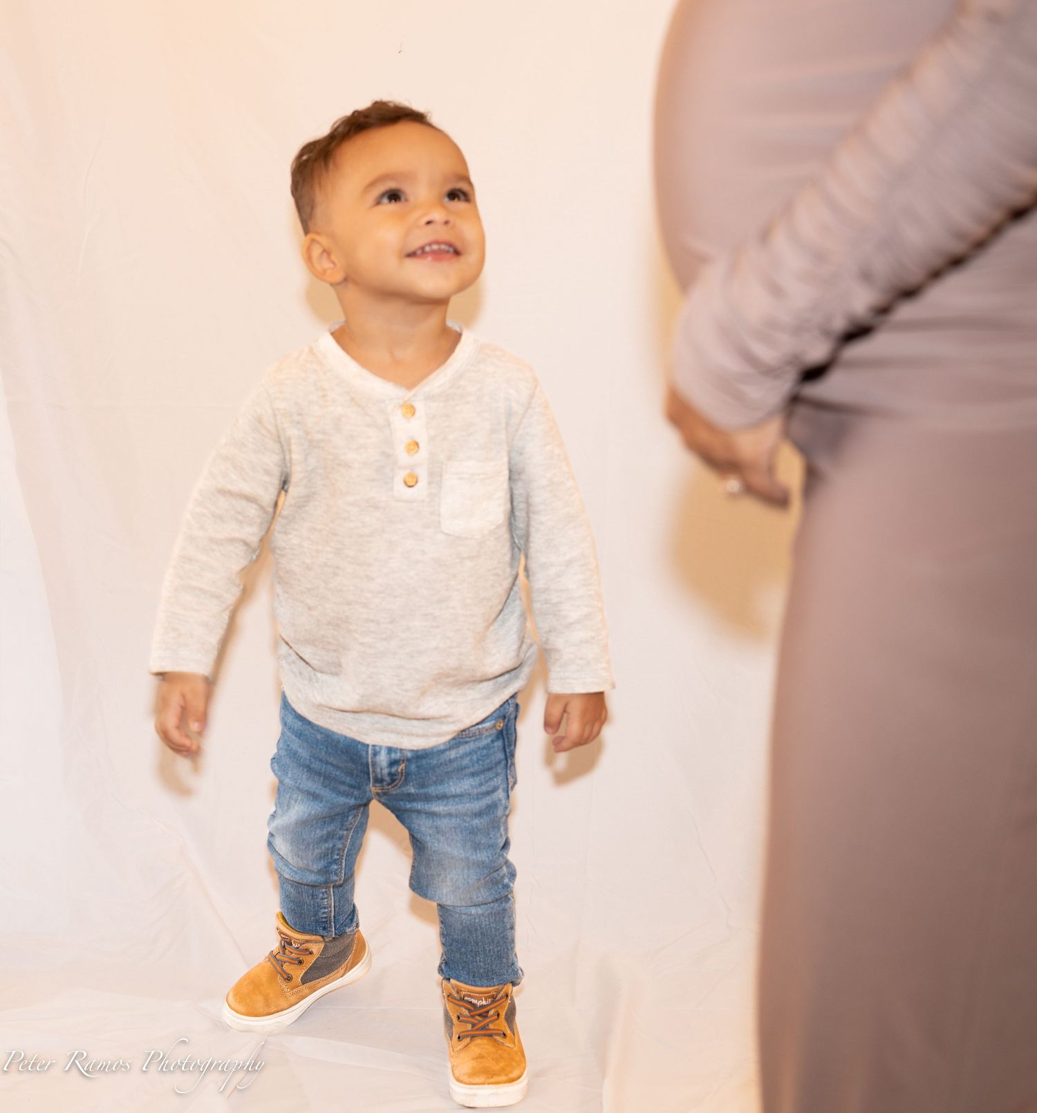 Young child in beige henley and jeans smiles while standing against a light backdrop.