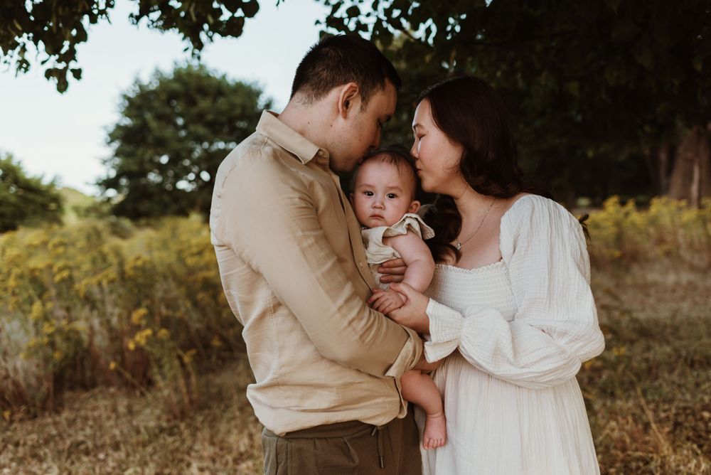 A tender family moment captured outdoors in warm sunlight with an infant being embraced by parents.