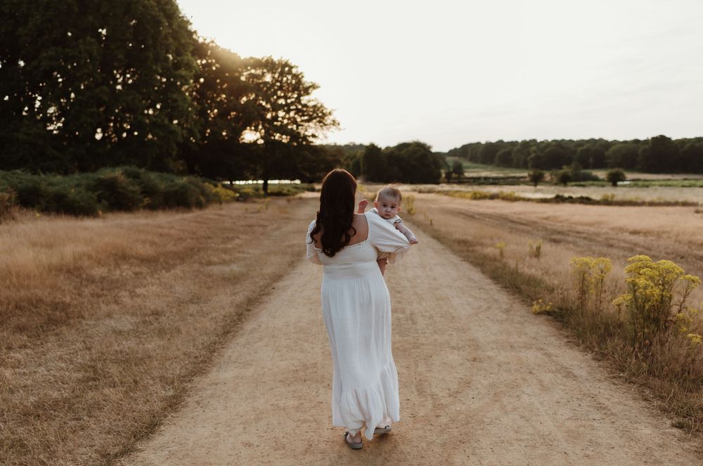 A person in a flowing white dress holds a baby while walking down a dirt path at sunset.