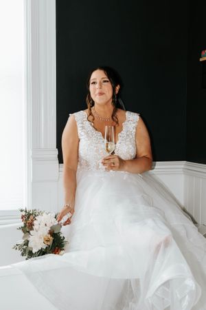 Bride in lace wedding gown holding champagne glass and floral bouquet seated by window.