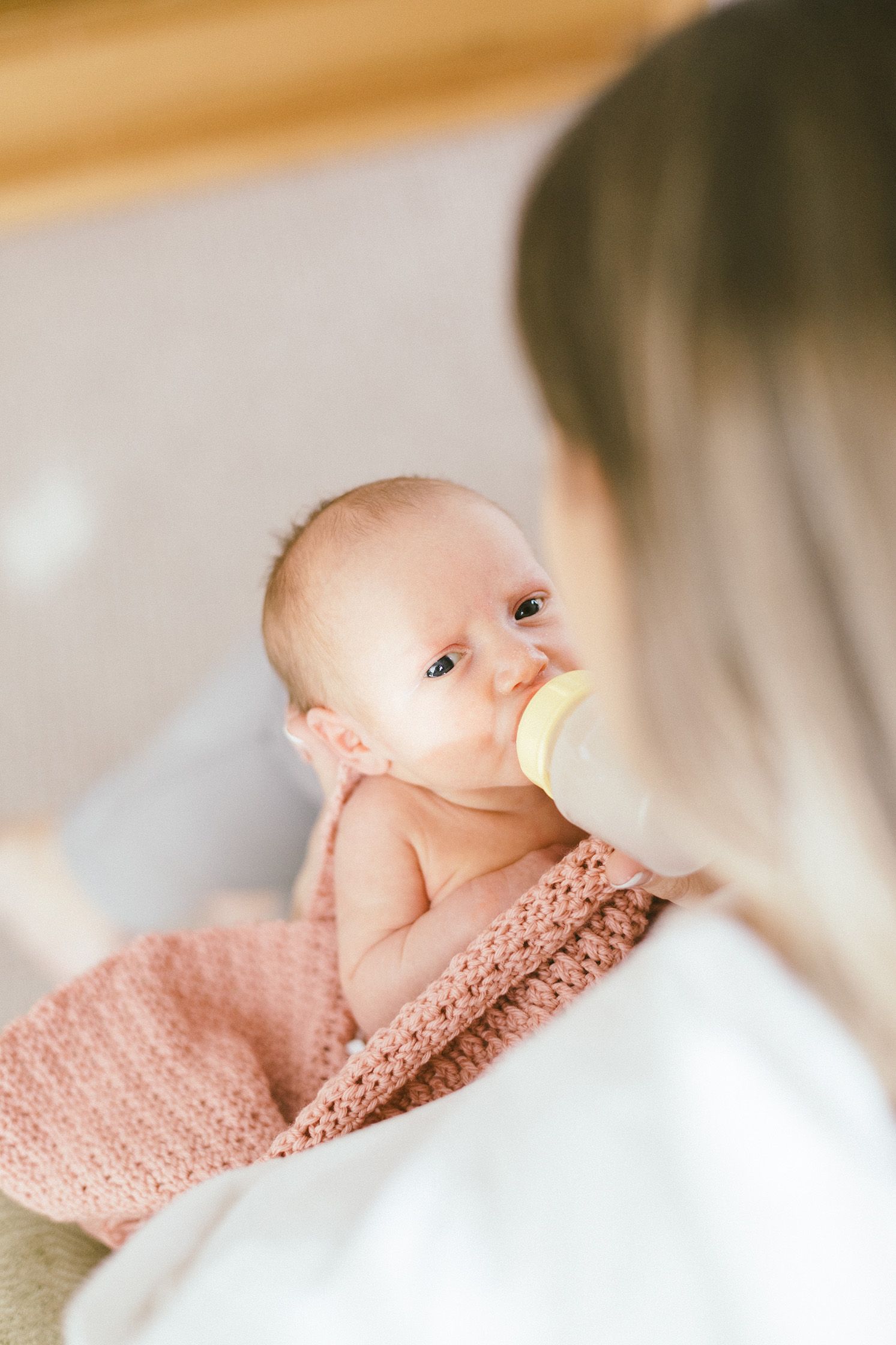 Baby in pink blanket drinking from bottle while being cradled in caregiver's arms.