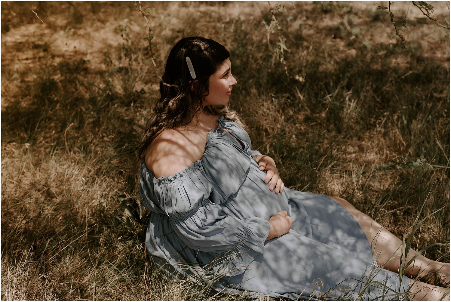 Ethereal maternity portrait with warm lighting and natural backdrop showing an off-shoulder grey gown.