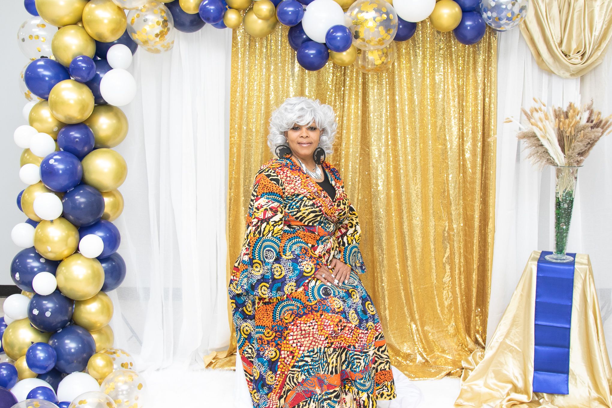 A woman in colorful dress poses against gold sequin backdrop with blue and gold balloon arch.
