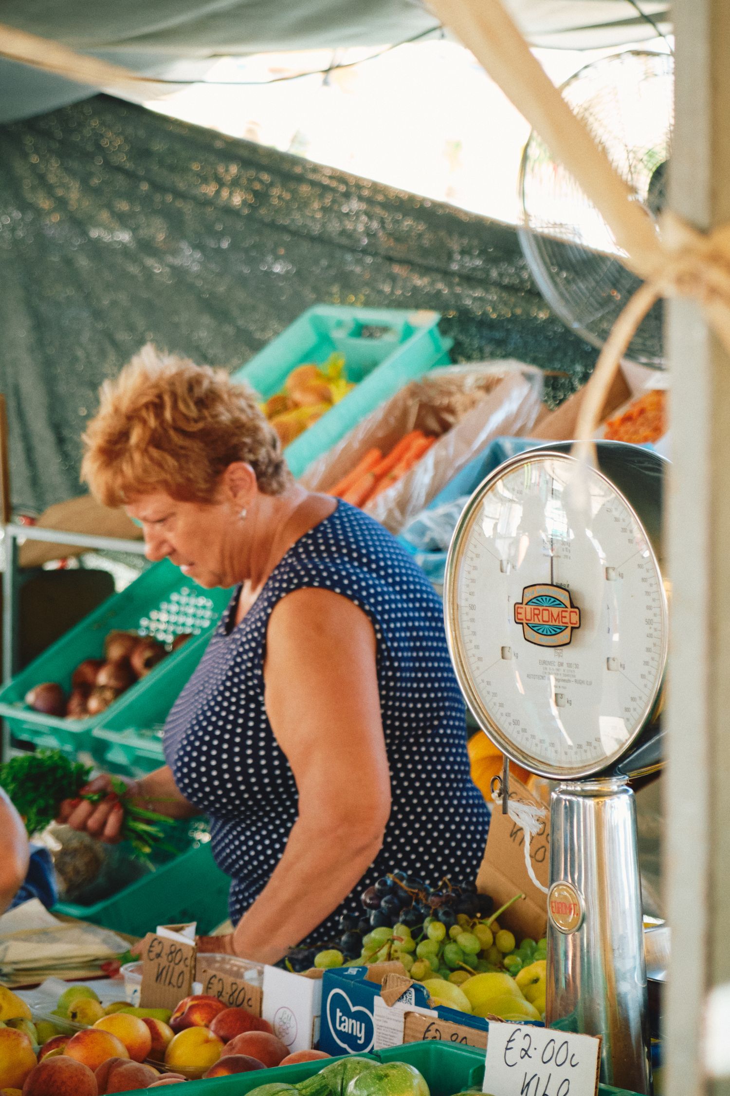 A vendor at a farmers market weighs fresh produce on a scale while surrounded by colorful fruits and vegetables.