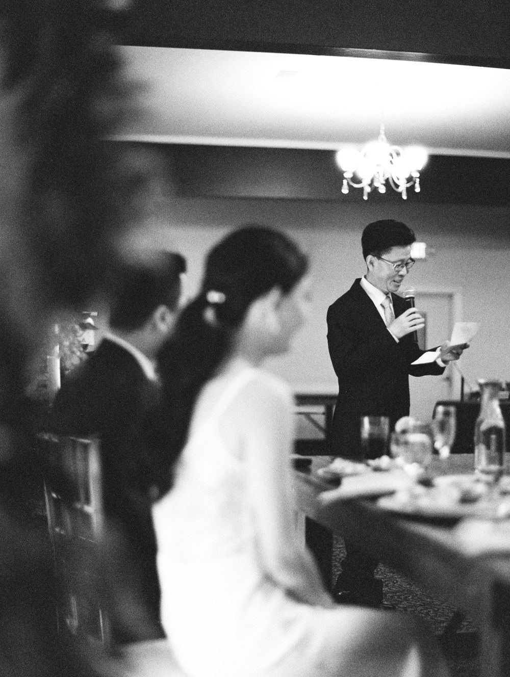 Wedding guests gather indoors at reception tables under chandelier lighting.