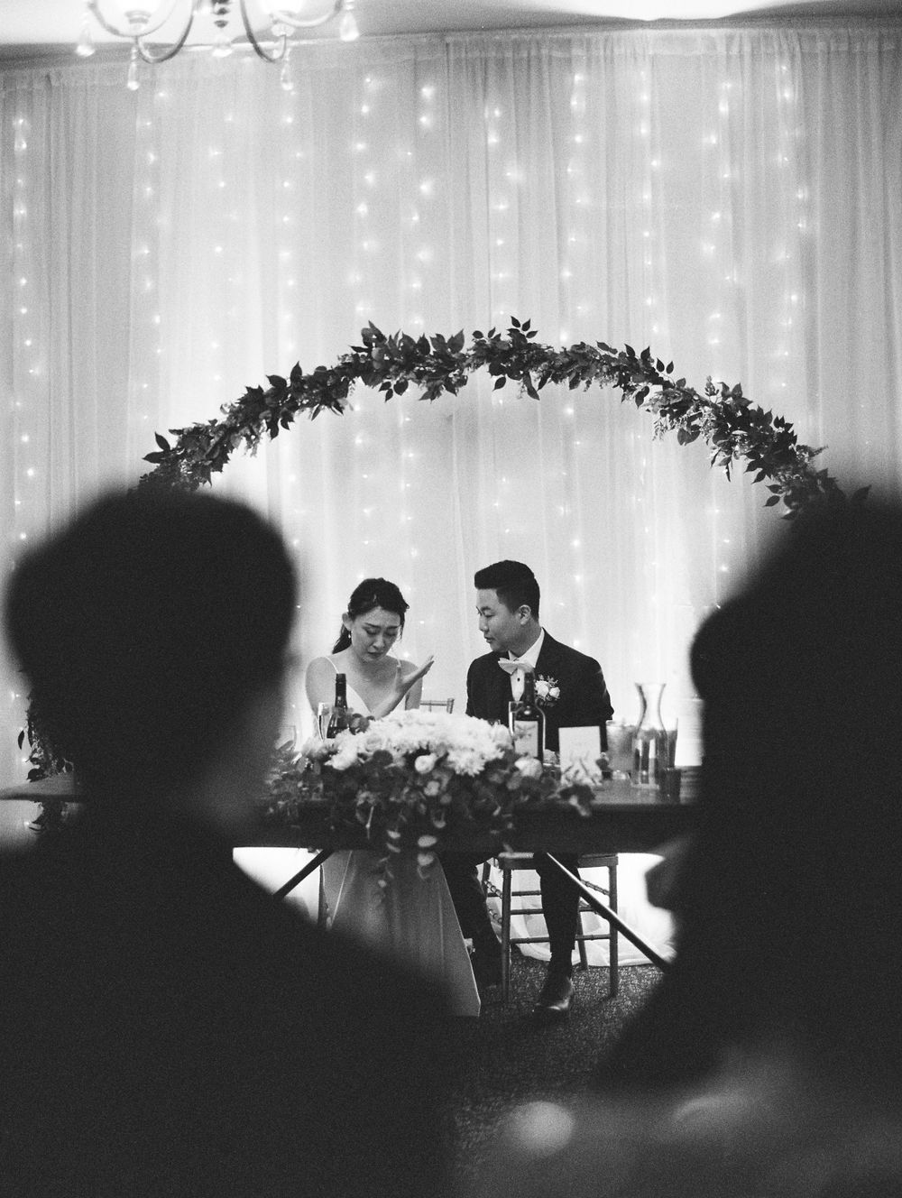 Elegant wedding reception scene with couple seated beneath floral arch and fairy light backdrop.
