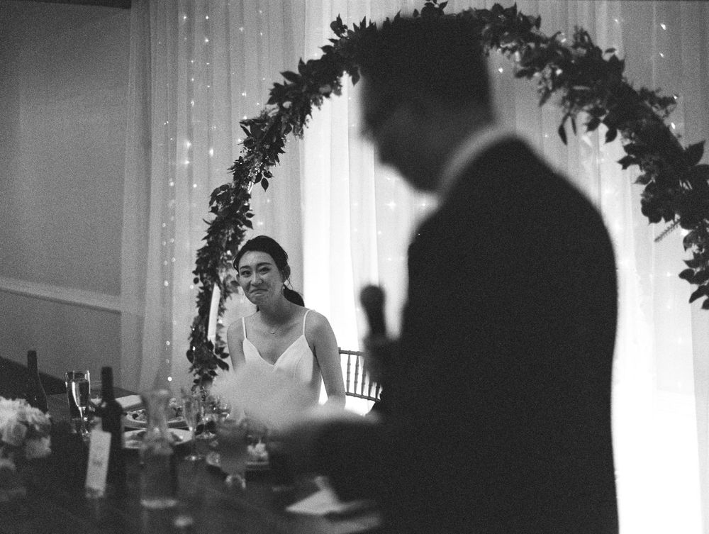 Black and white photo showing couple at reception head table under floral arch with sparkle lights in background.