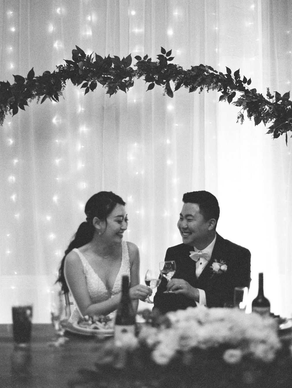Couple sharing intimate moment at reception table under decorative leaf arch and twinkle lights.