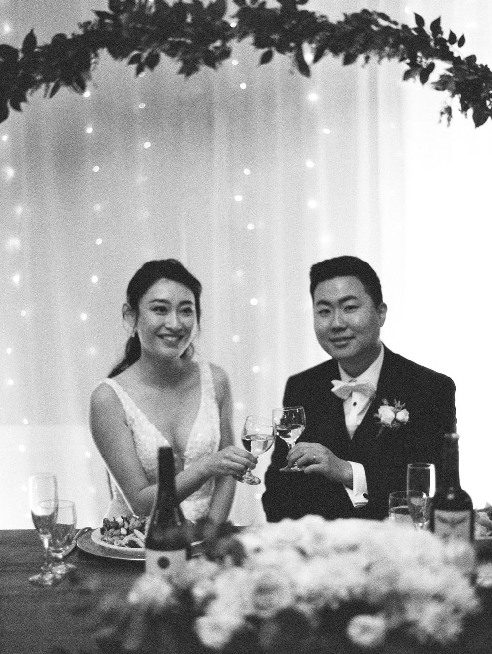 Couple toasts with wine glasses at wedding reception under floral arch and twinkling backdrop.