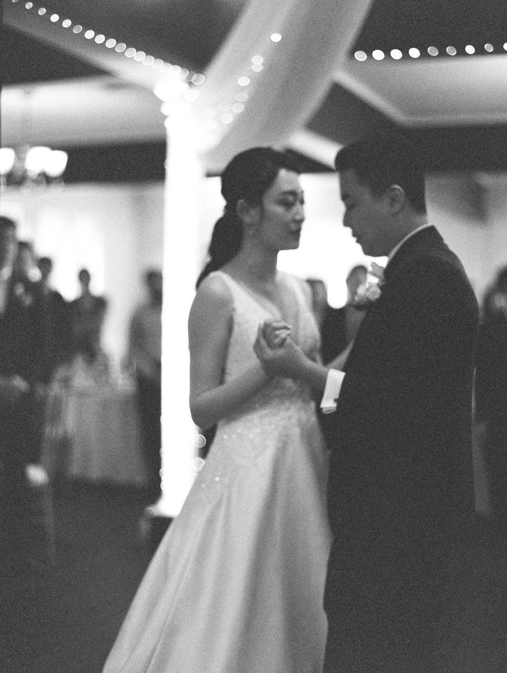 Intimate black and white photo of couple dancing beneath string lights at wedding reception.